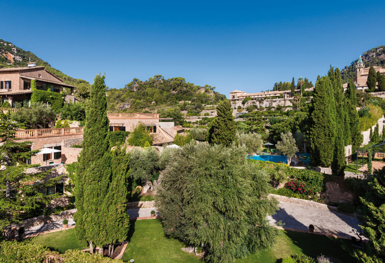 Aerial view of the hotel's stone terraces and pool set against olive groves, with Valldemossa's monastery on the hillside behind