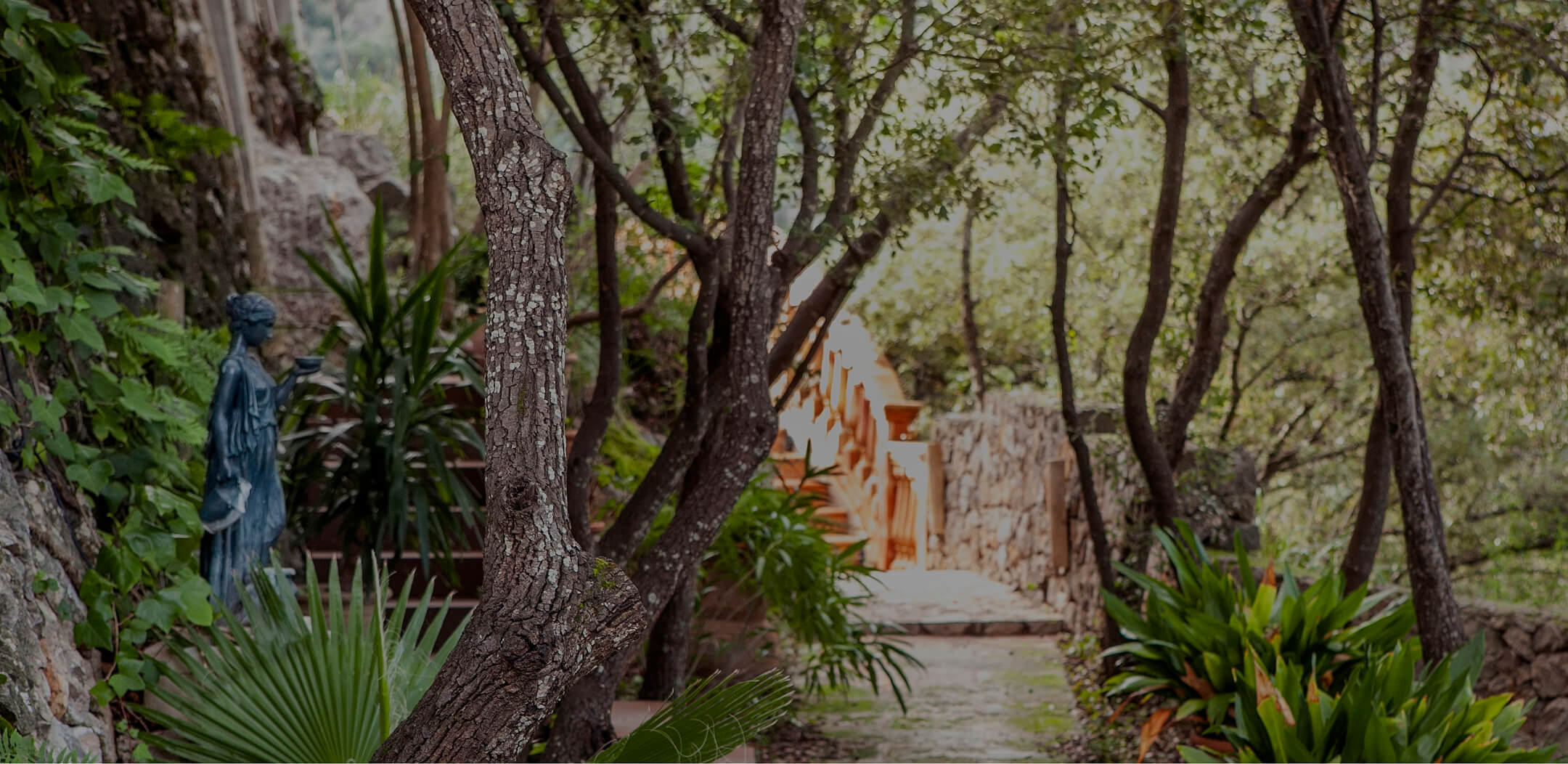Shaded garden path through trees leading to a stone archway, with a statue among the plantings