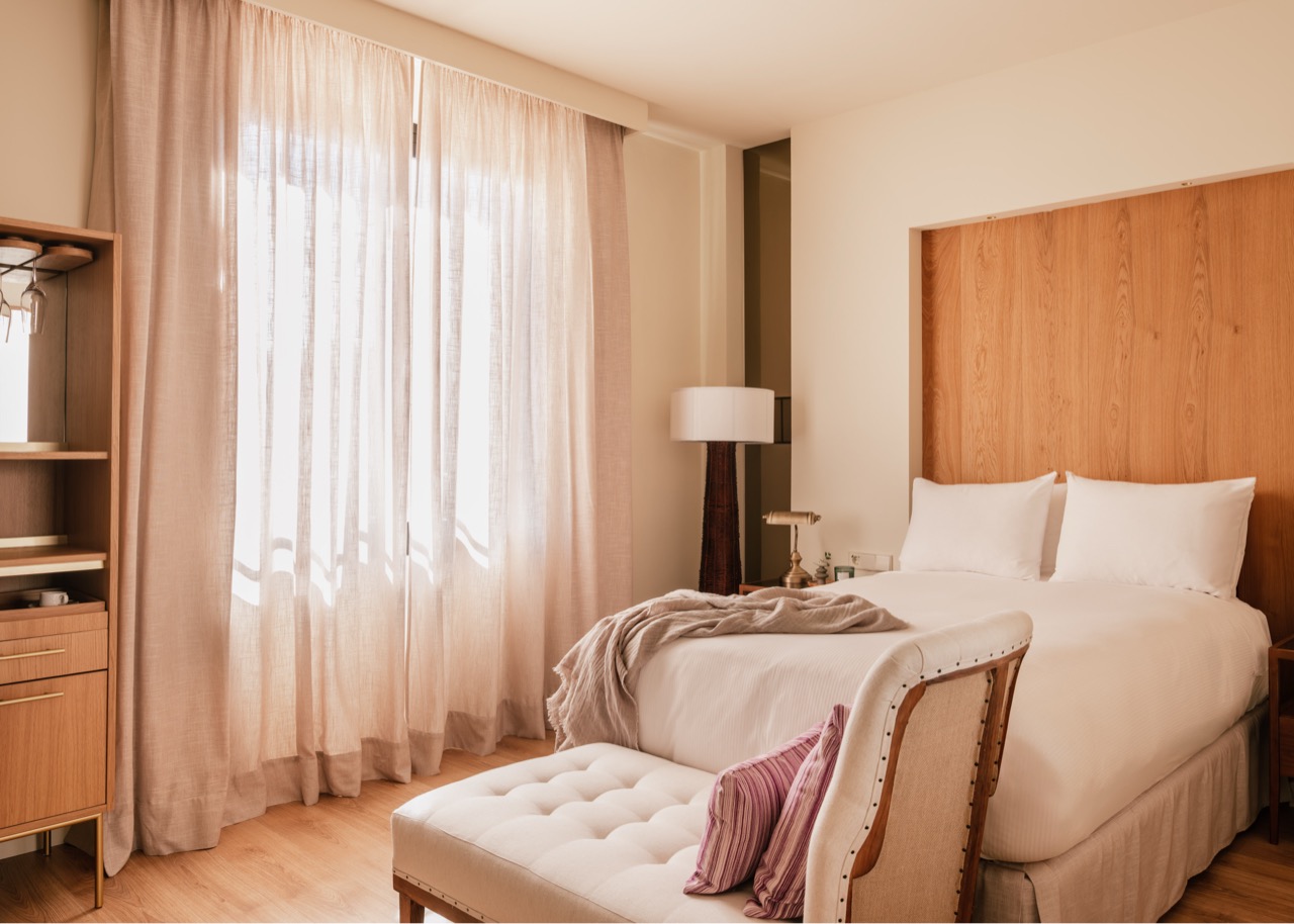 Guest room with wood headboard, sheer curtains and a tufted bench at the foot of the bed