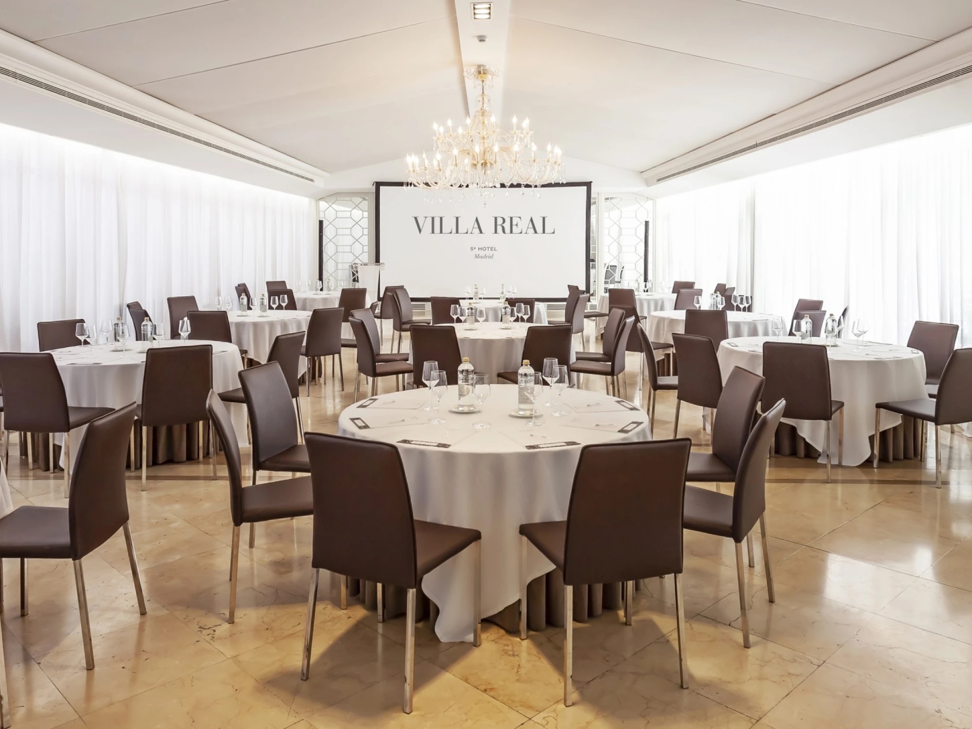 A function room set with round white-clothed tables and brown leather chairs beneath a chandelier, with a screen reading Villa Real at the far end.