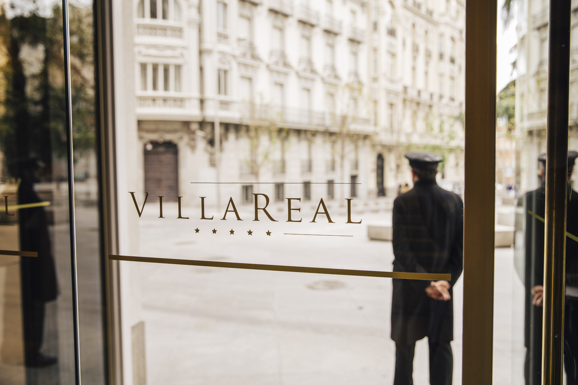 The glass entrance door with Villa Real lettered in gold and a uniformed doorman standing outside facing the square.