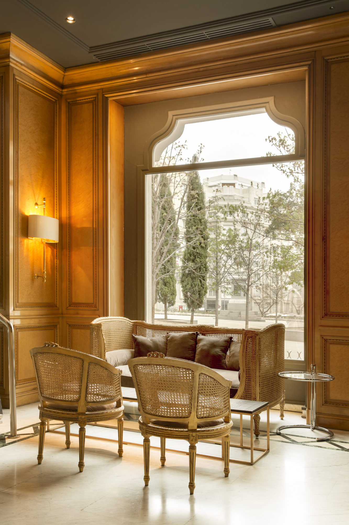 A lounge corner with a cane-backed sofa and two cane armchairs by a tall arched window, set against wood-panelled walls with a lit sconce.