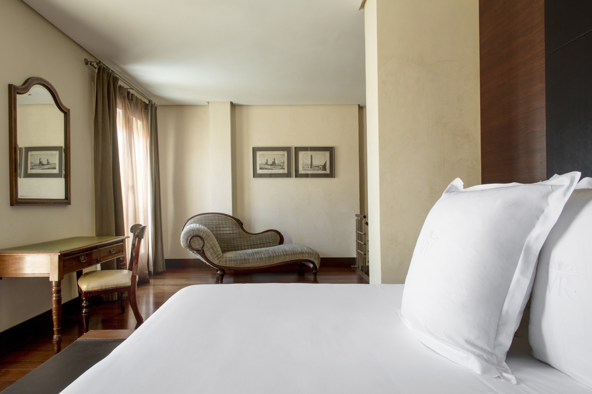 A guest room with a white-linen bed whose pillows are embroidered with a VR monogram, a mahogany writing desk and chair by a curtained window, a striped chaise longue and two framed prints on a pale wall.