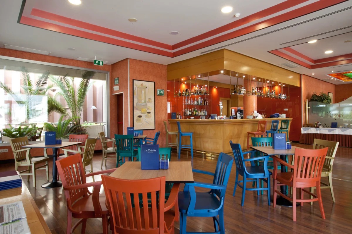 A colourful dining and bar area with a wooden bar counter, mismatched painted chairs, and table menus branded La Pirámide Restaurante.