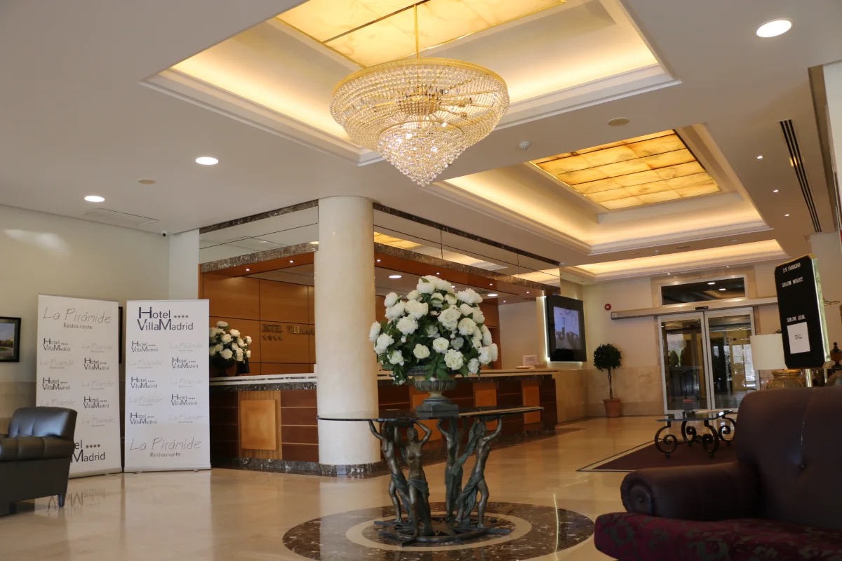 The hotel lobby with a crystal chandelier, a marble reception desk, and signage for both Hotel VillaMadrid and La Pirámide Restaurante.