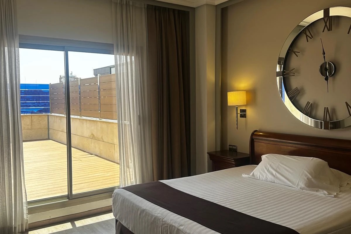 A guest room with a wooden headboard, an oversized round wall clock, and a sliding door opening onto a private terrace with a city view.