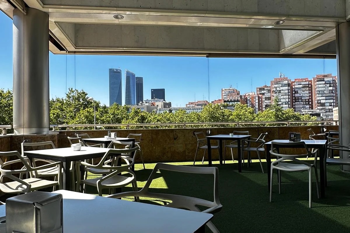 A covered terrace with tables and chairs on artificial turf, looking out over trees toward the Cuatro Torres skyscrapers.