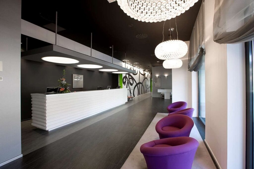 The hotel's white reception desk staffed behind computer terminals, with round pendant lights overhead and purple tub chairs along the corridor.