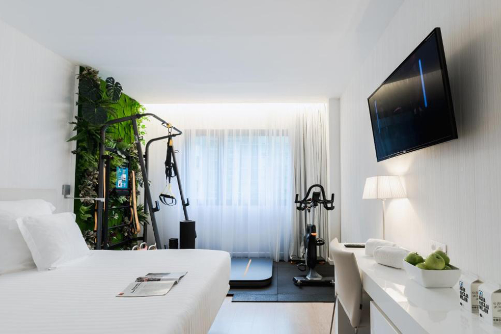 A guest room set up as the Fitness Room type, with a treadmill, spin bike and TRX straps beside the bed, a wall-mounted TV and a desk.