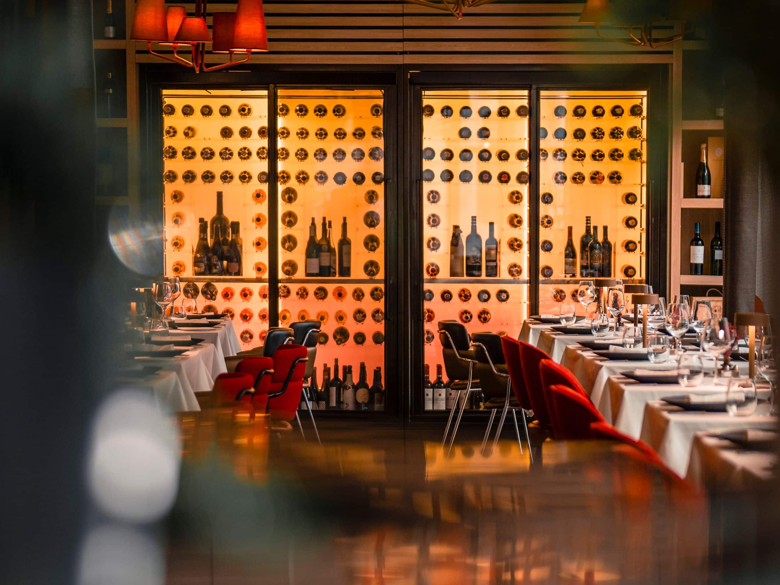 Dining room with white-clothed tables, red and black chairs and a backlit wine display wall.