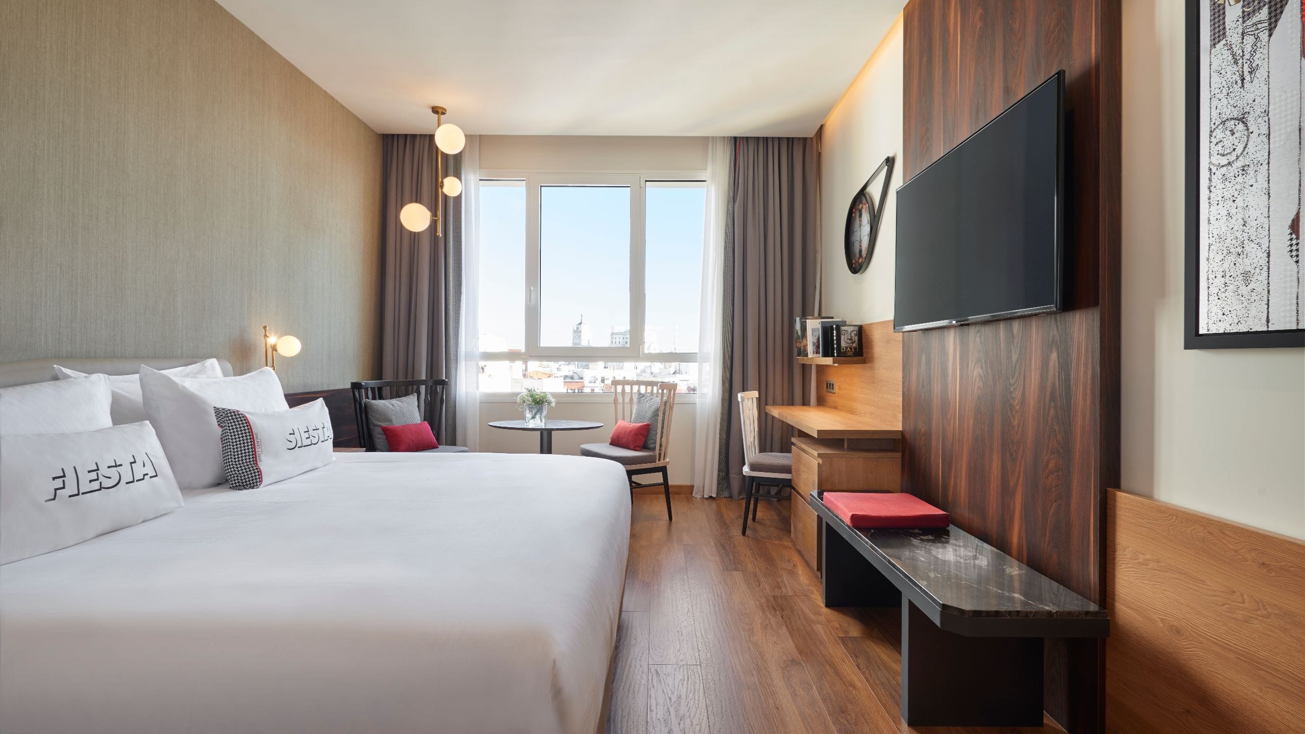 Guest room with a king bed, a wall-mounted TV on a dark wood panel and a window over city rooftops.
