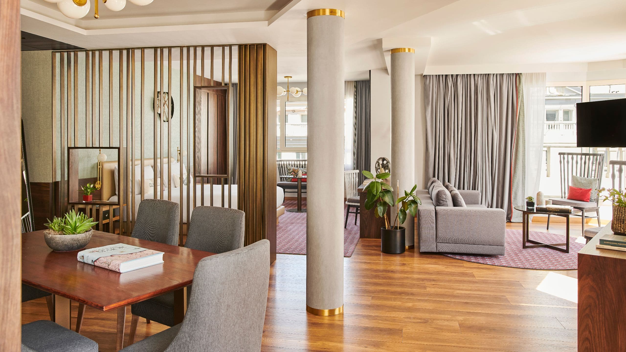 Open-plan suite with a dining table, lounge seating and a bedroom behind a brass slatted partition.