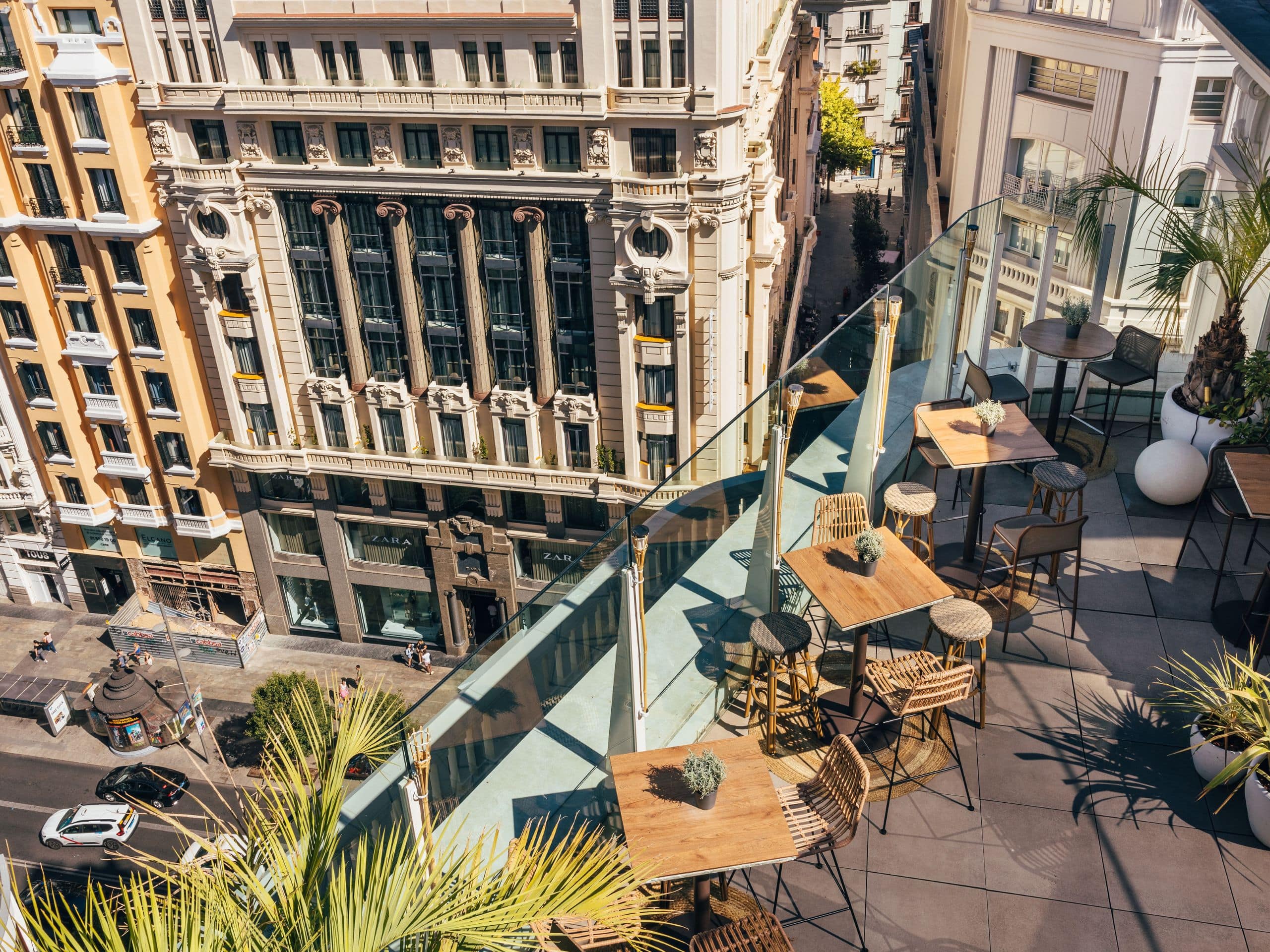 Rooftop terrace with high wooden tables and stools behind a glass balustrade, overlooking the ornate buildings across Gran Via.