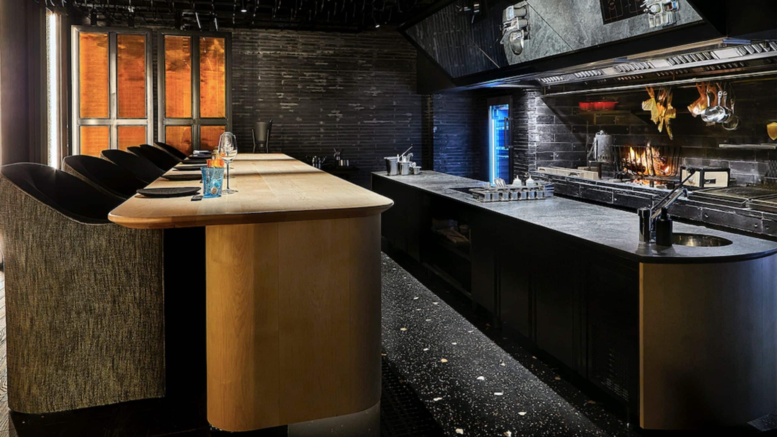 Counter seating at an open kitchen with a wood-fired grill and dark brick walls.
