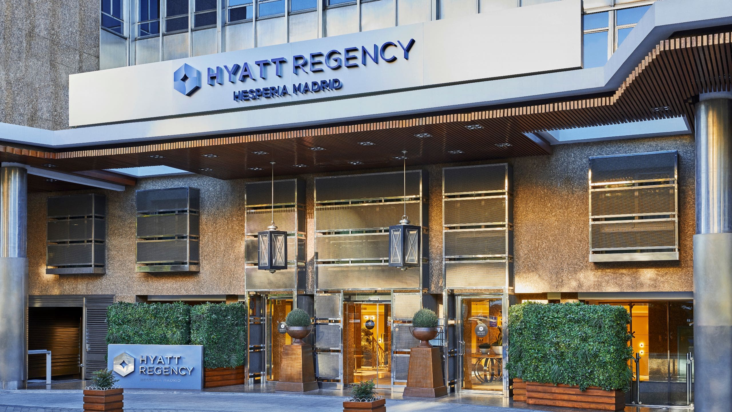 Street entrance beneath a Hyatt Regency Hesperia Madrid sign, flanked by green hedges and hanging lanterns.
