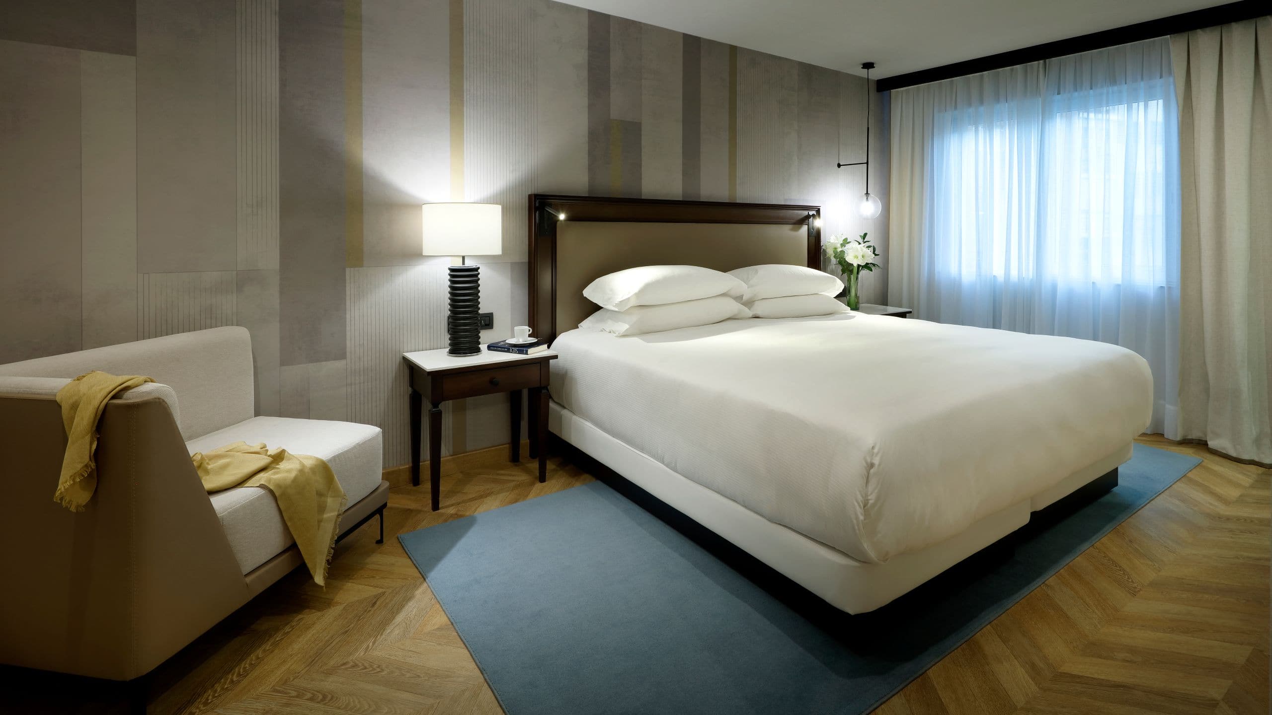 Guest room with a king bed, an upholstered headboard, a bedside lamp and a chaise longue by the window.