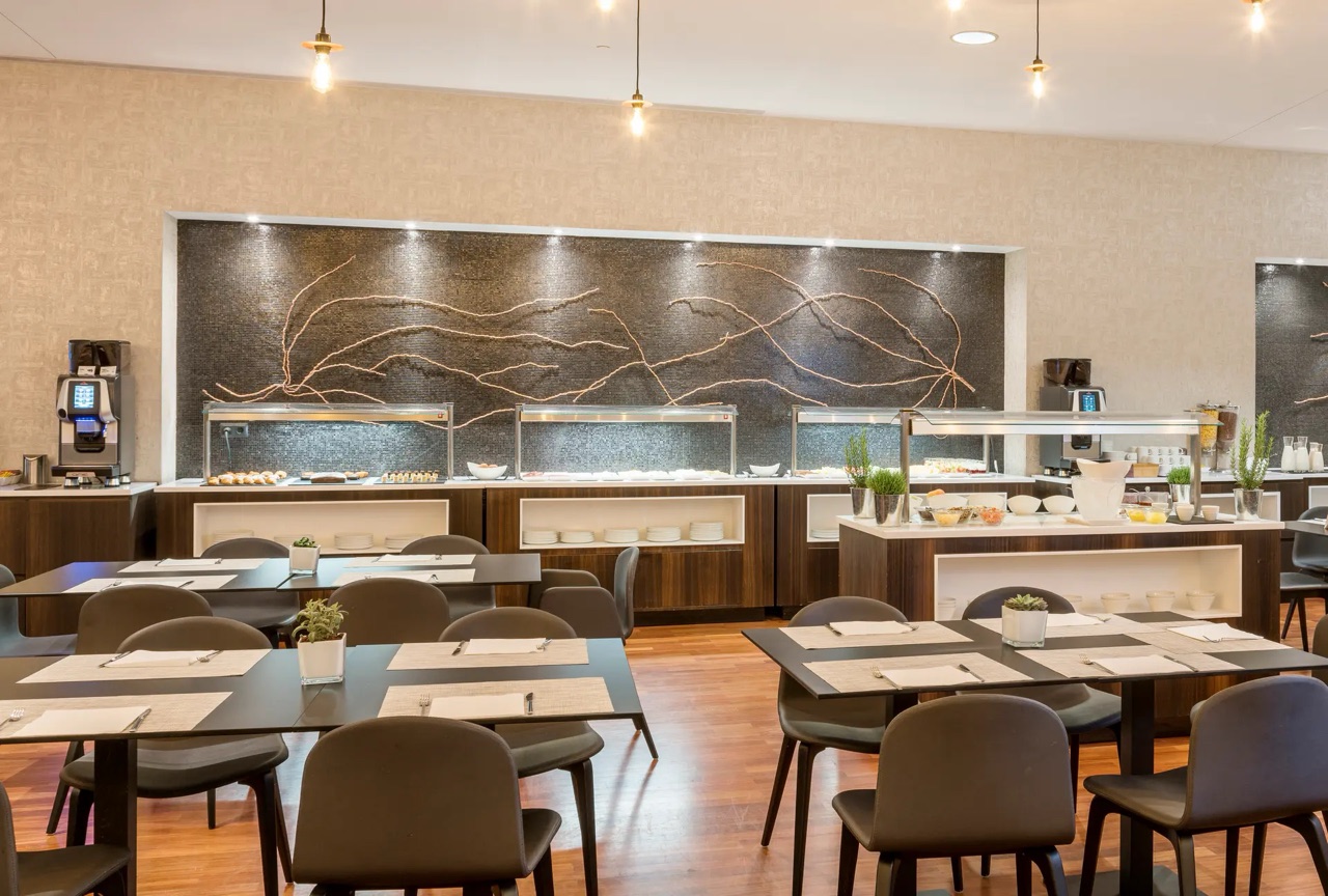 A dining room with dark wood tables set with placemats, facing a buffet counter with chafing dishes and a mosaic wall behind it.