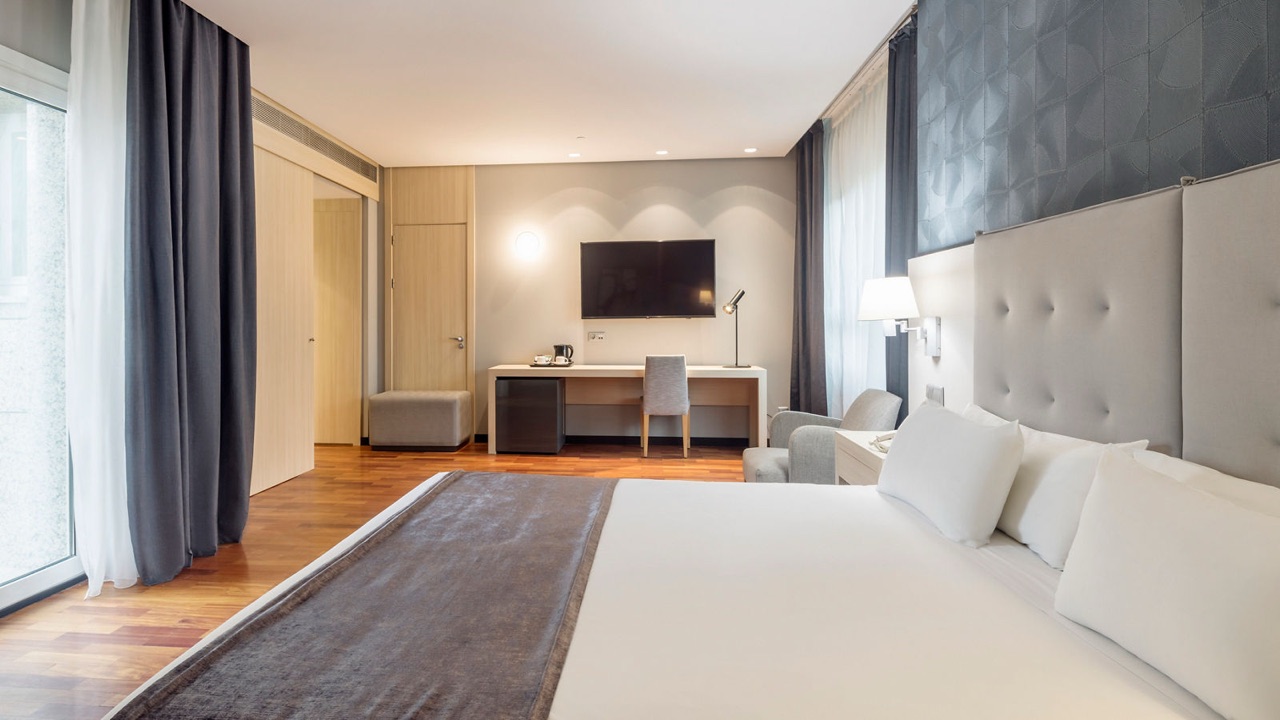 A hotel room with a grey upholstered headboard, white bedding, a desk with a kettle, and a wall-mounted TV.