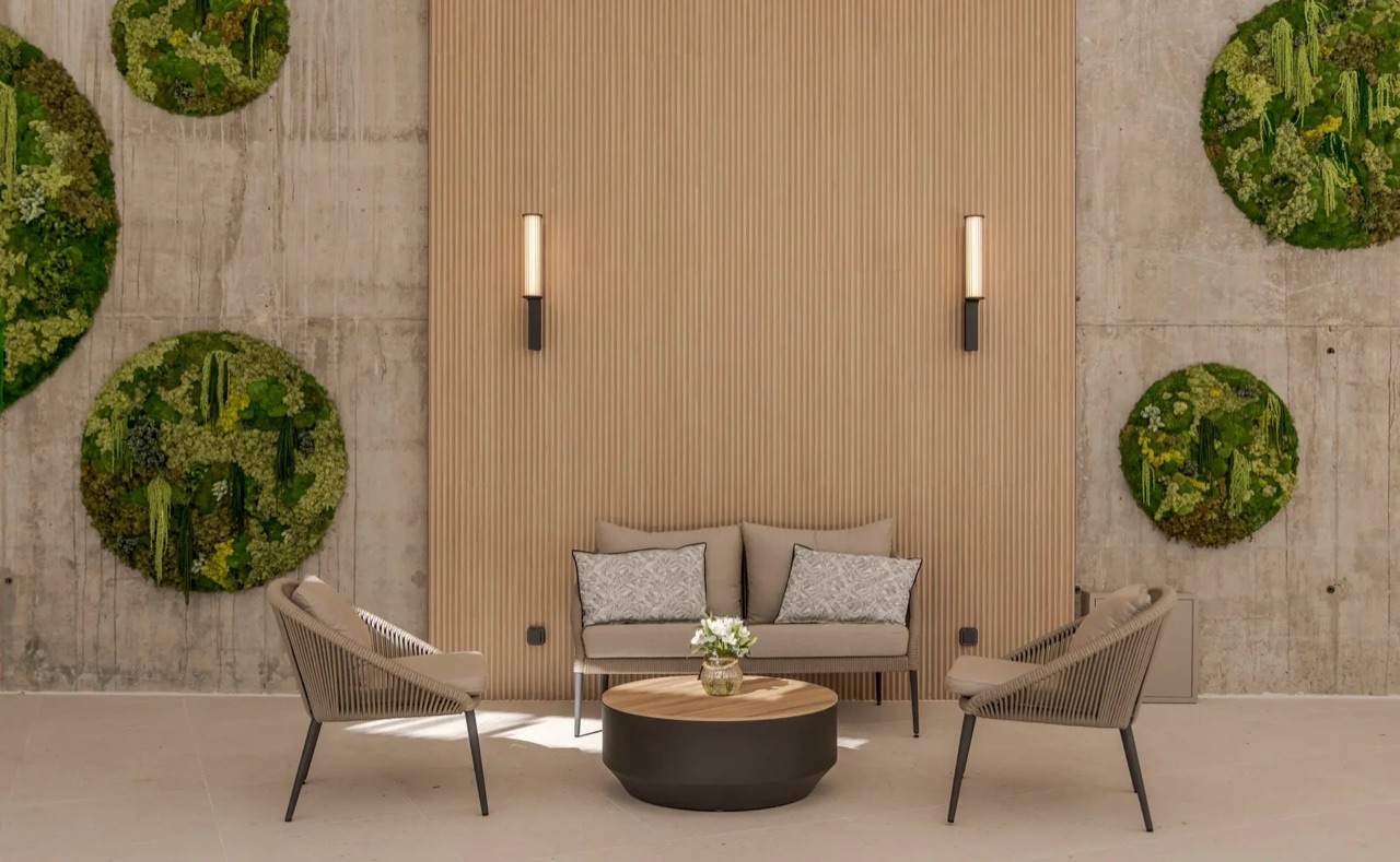 An outdoor lounge corner with woven armchairs, a sofa, a round coffee table, and circular moss wall panels against a concrete wall.