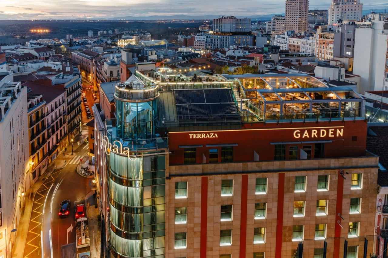 An aerial dusk view of the rooftop terrace at Inhala Hotel Garden, showing the glass-enclosed dining area, an open-air bar and Madrid's skyline behind it.