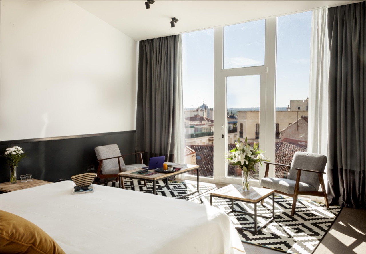 A hotel room with a bed dressed in white and green linens, next to floor-to-ceiling windows opening onto a balcony with rooftop views over Madrid.