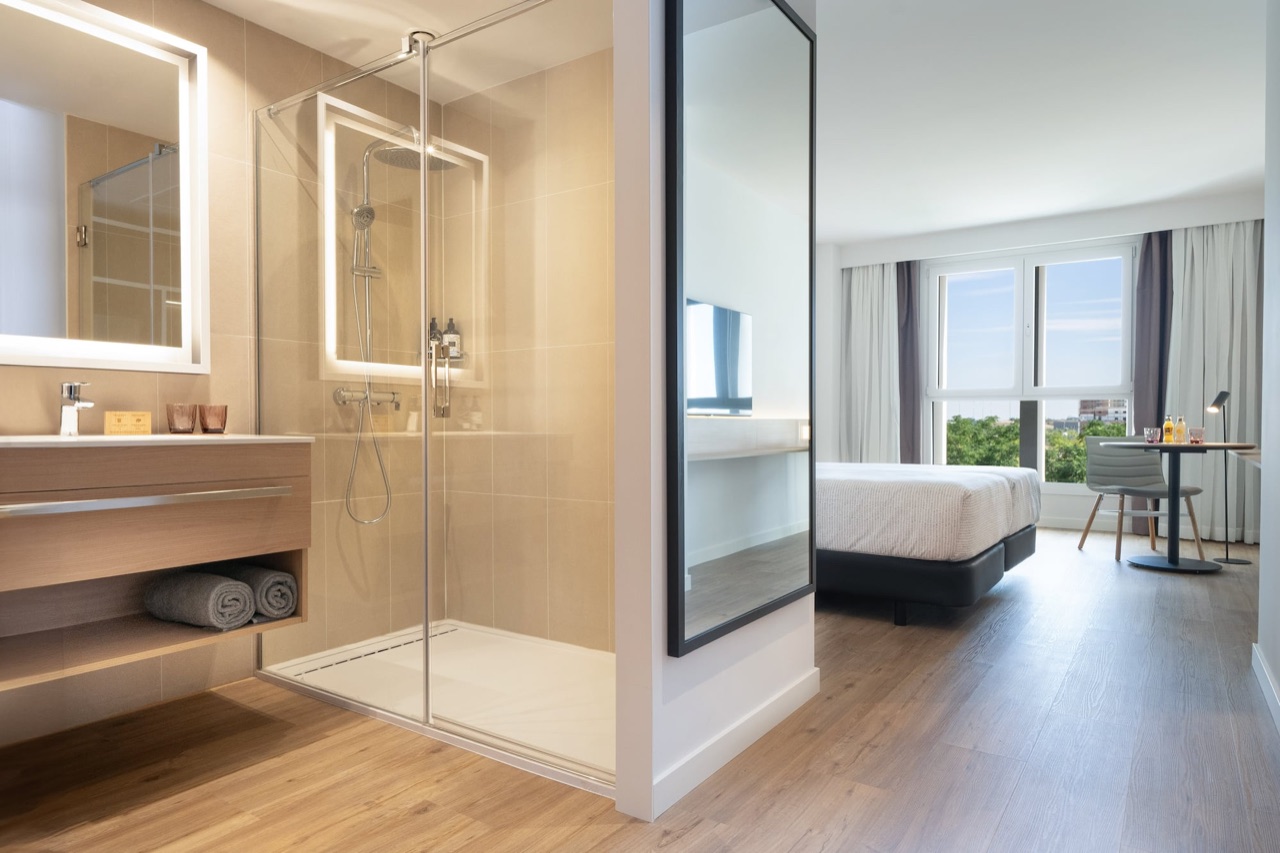 A hotel bathroom with a floating wood vanity, backlit mirror, rolled towels, and a glass-walled shower with a rain showerhead.