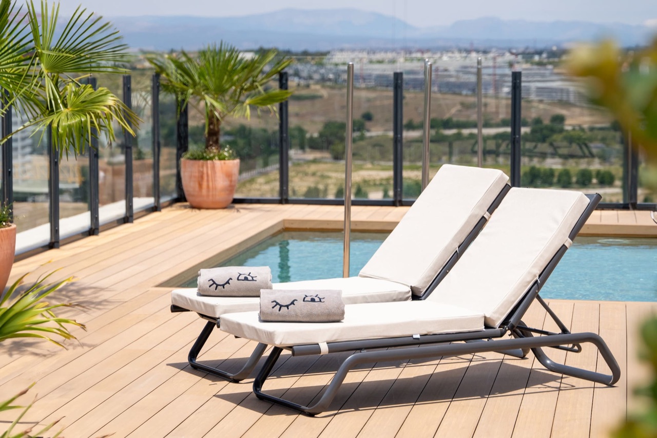 Two sun loungers with rolled towels beside the rooftop pool, with glass railings and a view over the surrounding Valdebebas area.