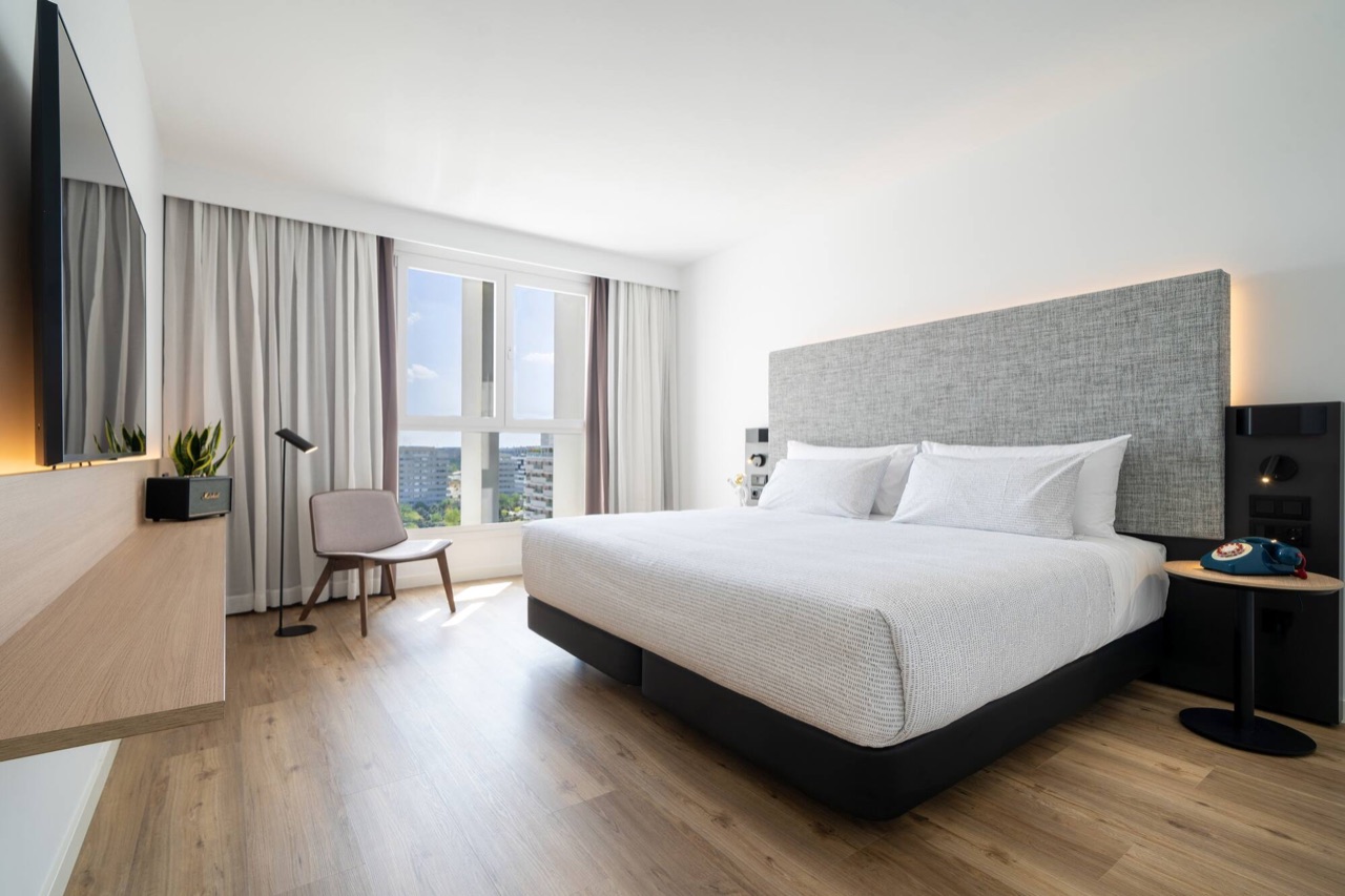 A hotel room with a grey upholstered headboard, white bedding, a Marshall speaker and a wall-mounted TV, with a city view through the window.
