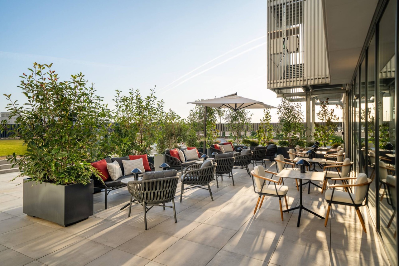 An outdoor terrace with woven armchairs, cushioned sofas, cafe tables and a large umbrella, surrounded by potted greenery.
