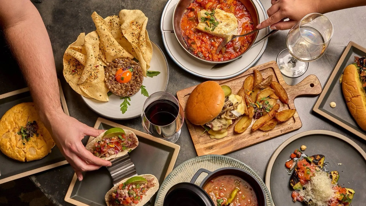 An overhead shot of a shared table at Antigua Embajada set with tapas plates, a burger, fried potatoes and glasses of wine.
