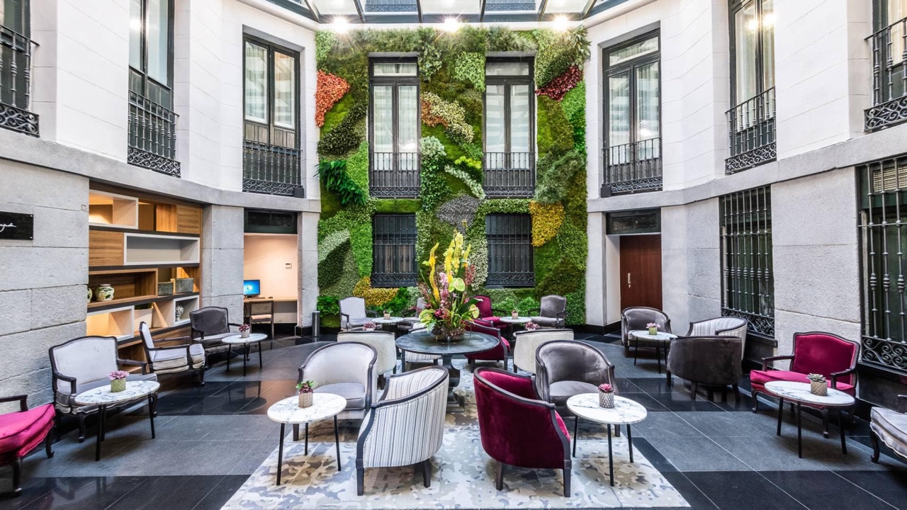 A glass-roofed courtyard lounge with velvet armchairs and marble side tables in front of a tall vertical garden wall covering the far facade.