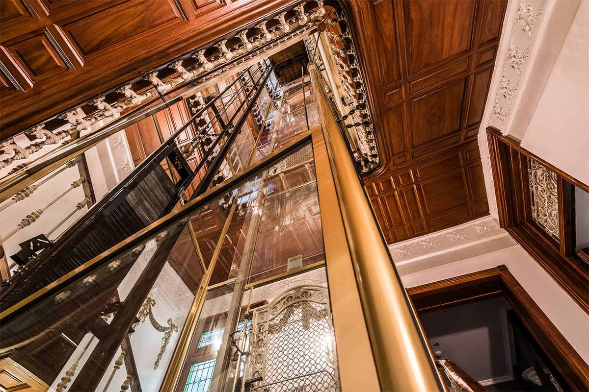 A view straight up an ornate wood-panelled stairwell with a brass-trimmed glass lift shaft at its center.