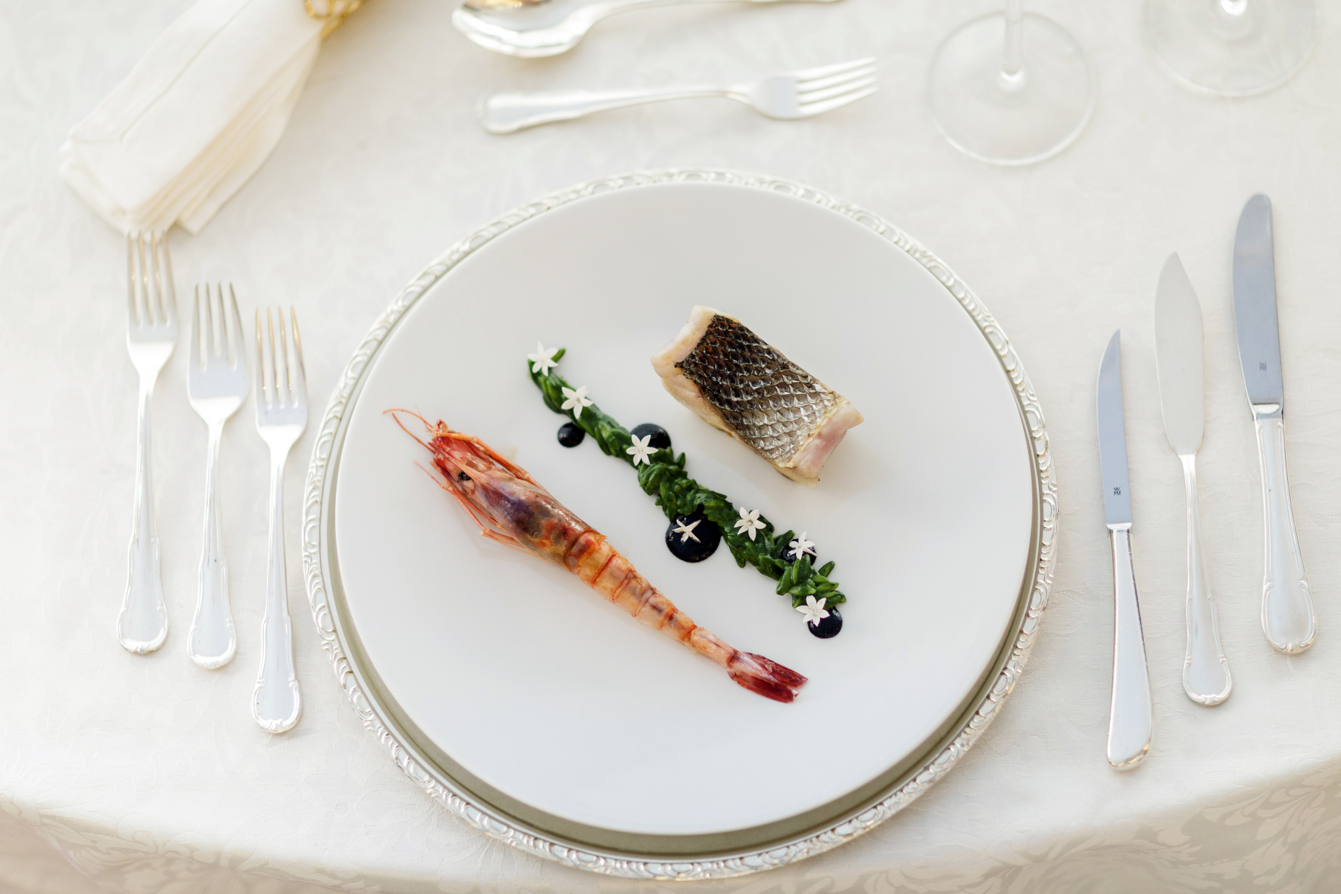 A white plate with a whole red prawn, a skin-on fish fillet and a line of sea greens dotted with small white flowers, on a formally laid table.