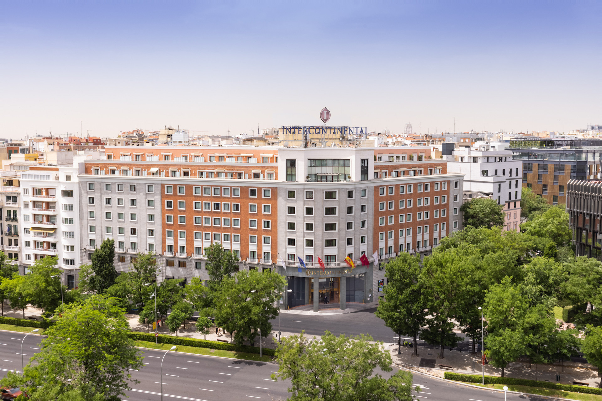 The hotel's brick and pale-stone facade seen from across a tree-lined avenue, with a rooftop InterContinental sign and a row of flags above the street-level entrance canopy.
