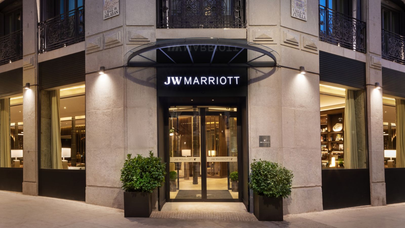 The corner entrance at street level: glass doors under a black canopy lettered JW Marriott, flanked by potted shrubs, with Calle de Arlaban and Calle de Sevilla tile plaques on the stone facade.