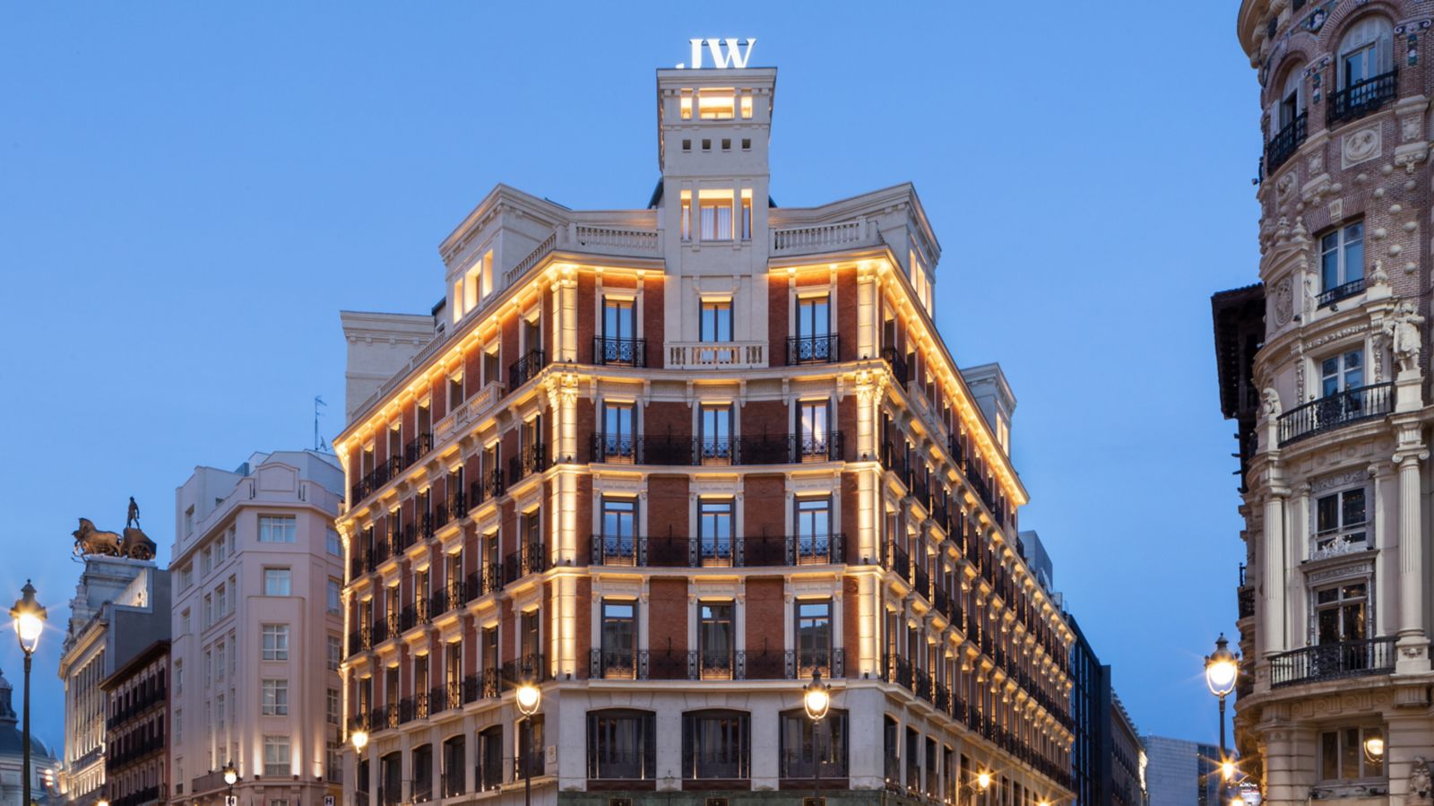 The hotel's corner facade at dusk: a brick and stone building with wrought-iron balconies, warm lighting along the cornices and an illuminated JW sign on the rooftop tower.