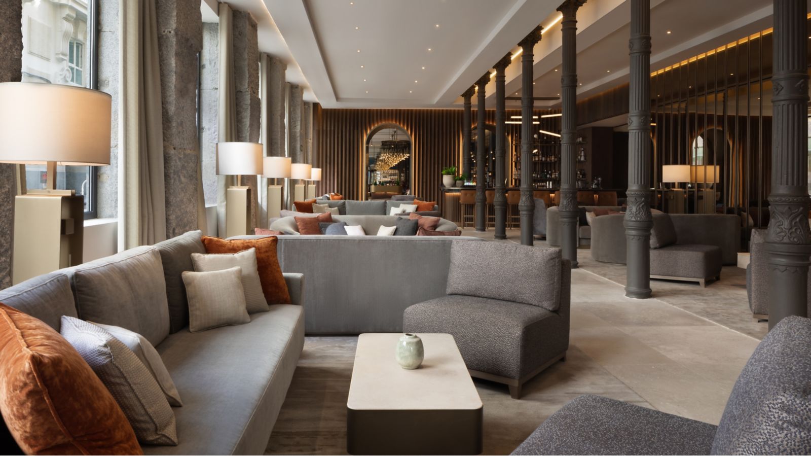 The lobby lounge: grey and taupe sofas with rust cushions around a low stone table, rows of slender cast-iron columns and a backlit bar at the far end.