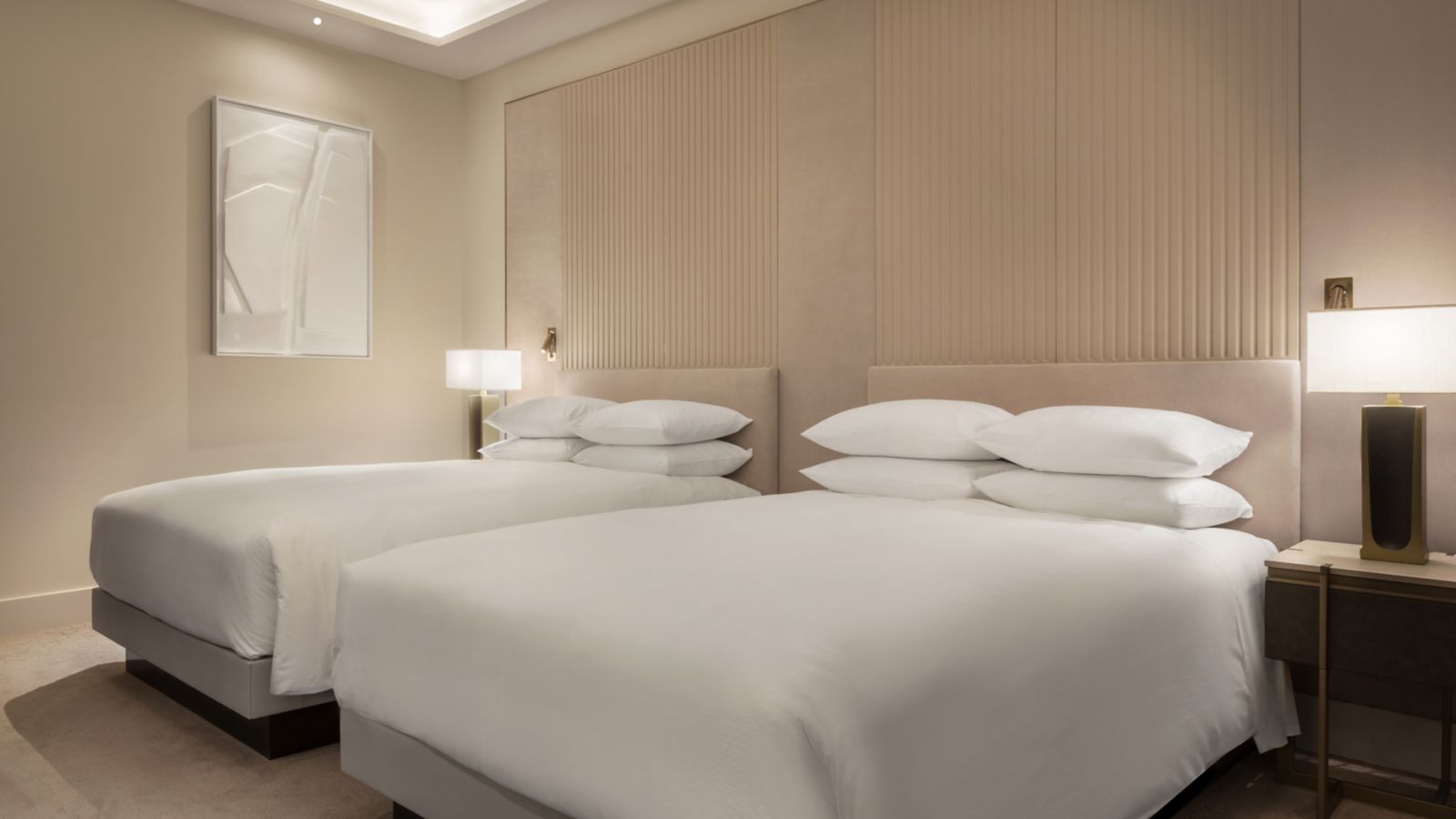 A twin room with two white-dressed beds against padded headboard panels, table lamps on either side and a framed white relief artwork on the wall.