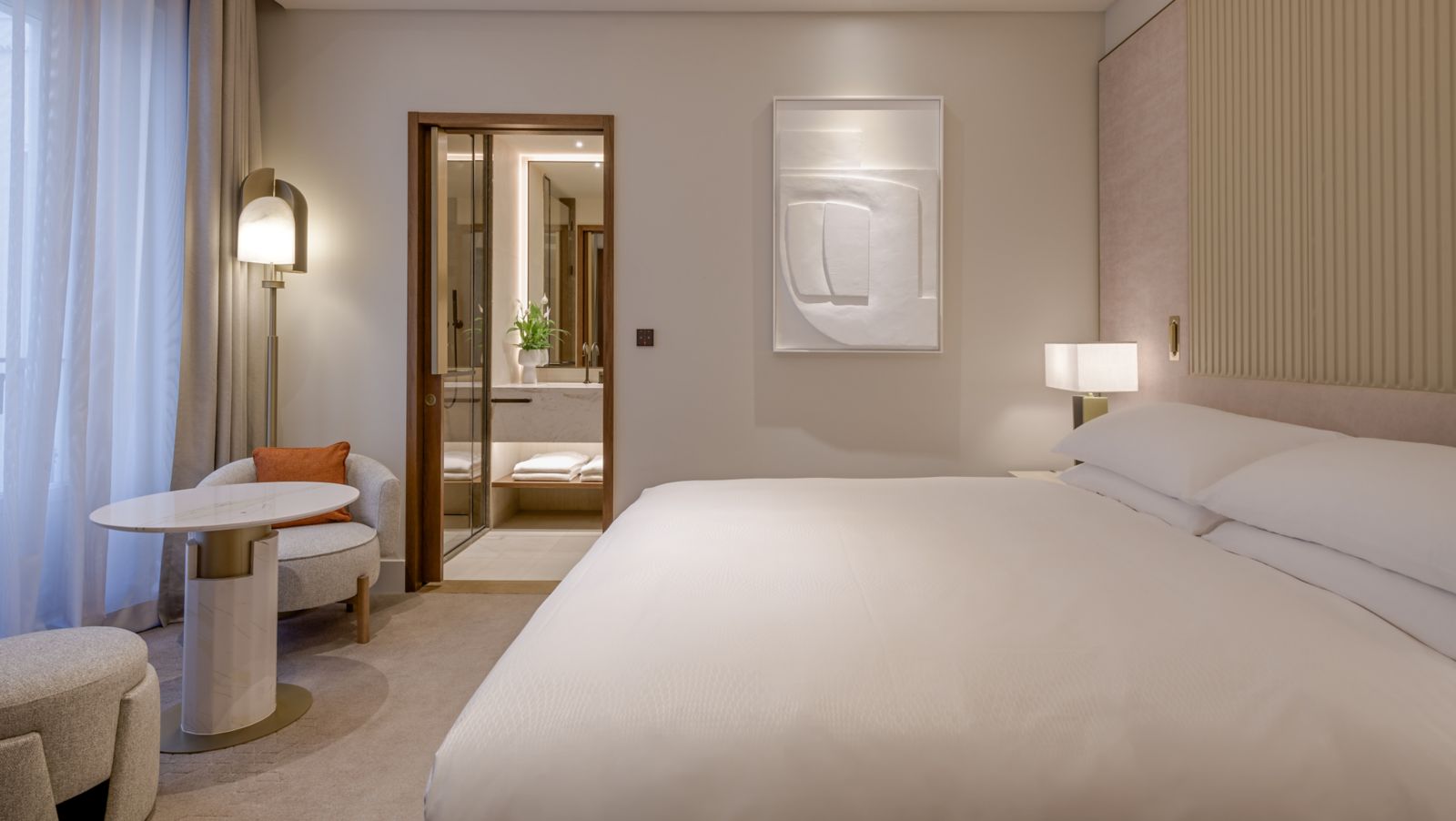 A guest room with a white-dressed king bed against a ribbed headboard wall, an armchair and marble side table by the curtained window, and a view into the marble bathroom.