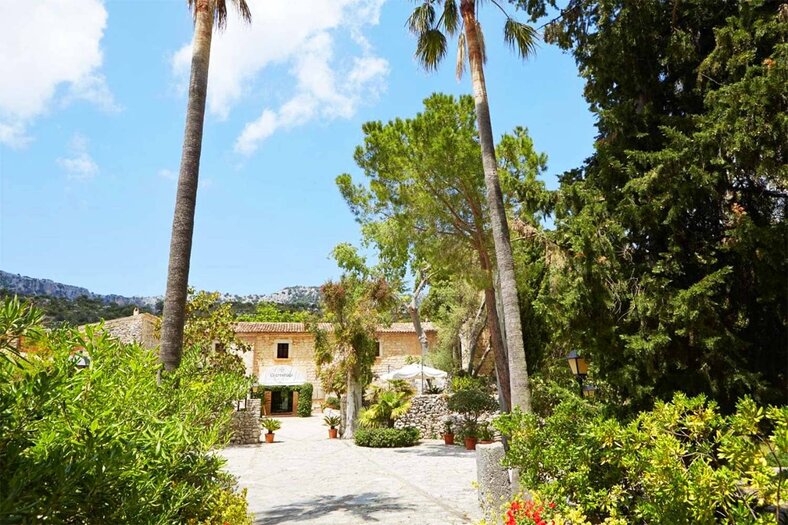 Stone facade of the former monastery, framed by tall palms at the end of a gravel entrance path