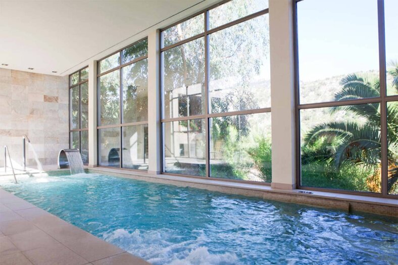 Indoor spa pool with a stone waterfall feature and floor-to-ceiling windows looking onto palms and hills