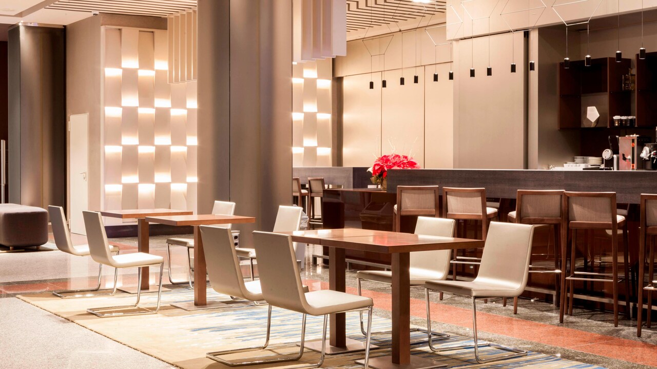 A hotel bar and lounge area with high stools along the counter and small tables with white chairs set on a patterned rug.
