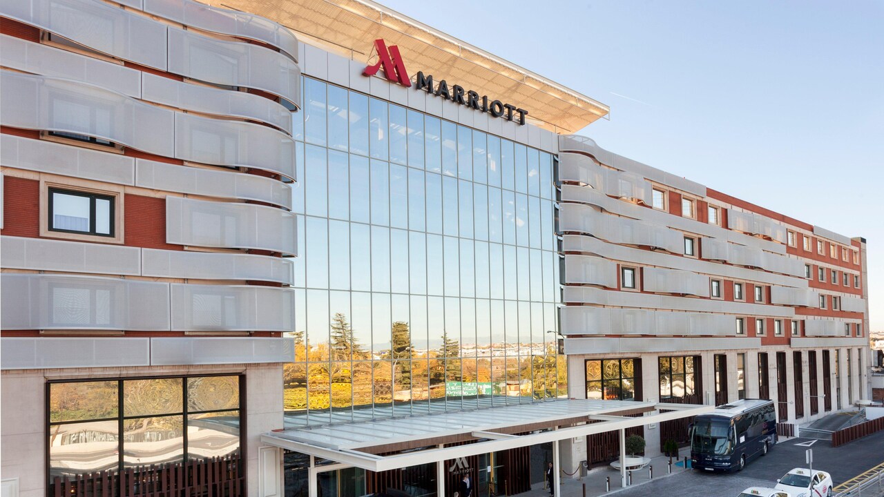 The hotel's glass and terracotta-brick exterior in daylight, with a Marriott sign on the rooftop and a coach parked at the covered entrance.