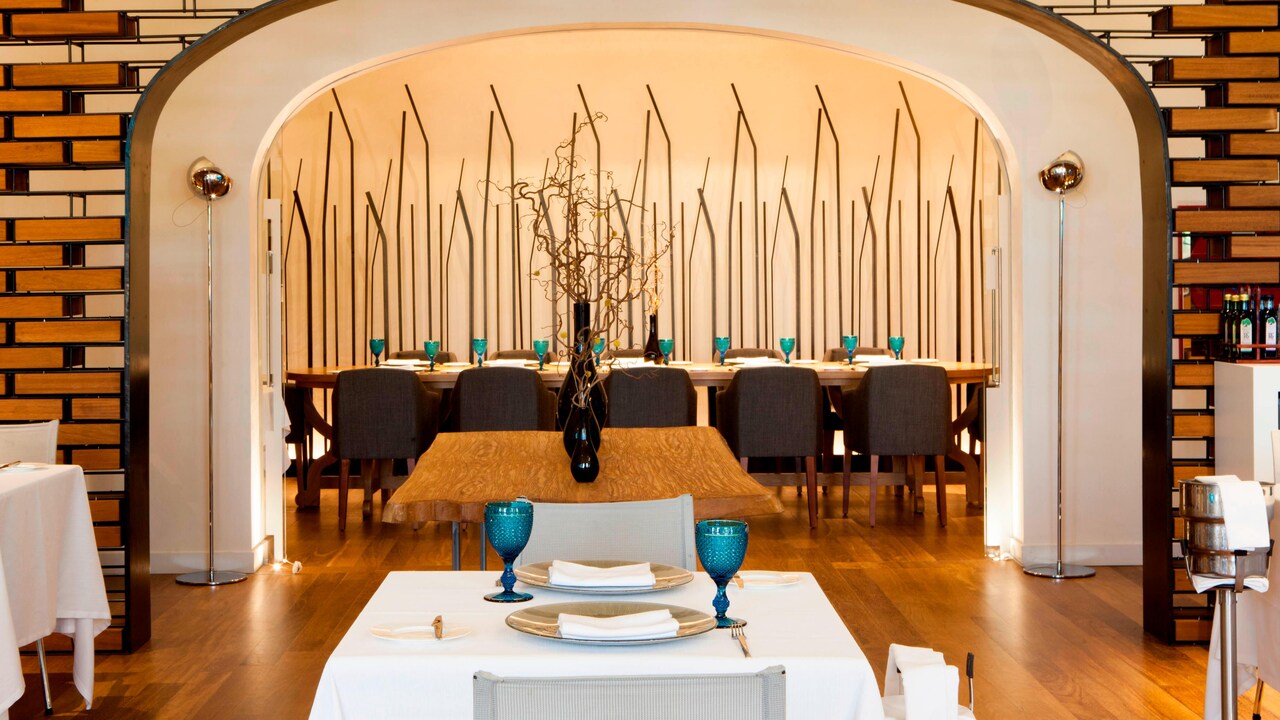A hotel dining room seen through an arched doorway, with a long communal table set with blue glassware and a smaller table set for two in the foreground.