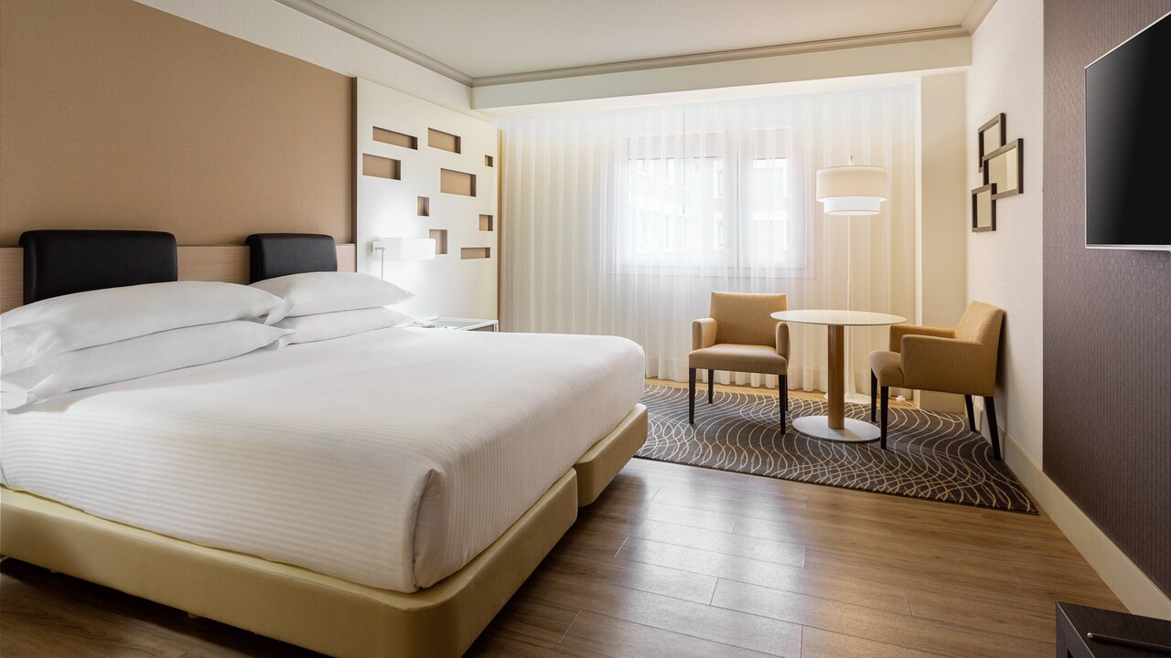 A hotel room with a king bed with a black leather headboard, and a small seating area with two armchairs and a round table by the window.