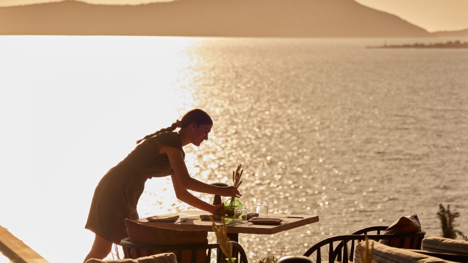 Waterfront dining terrace at Mandarin Oriental Punta Negra at sunset, with a staff member setting a table as golden light reflects off the sea