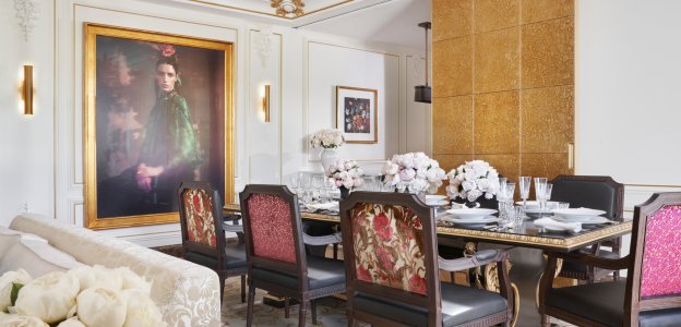 A dining room with a gold-edged table set for six with glassware and pink floral centerpieces, red-patterned chairs and a large gilt-framed portrait.