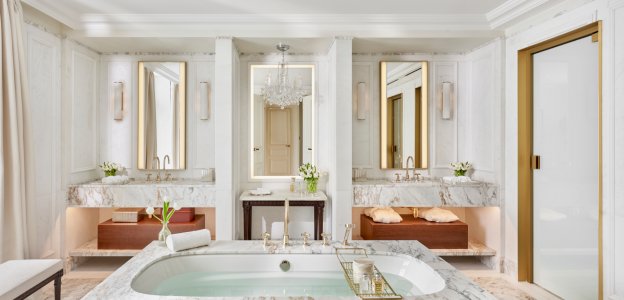A marble bathroom with a freestanding tub, twin vanities beneath lit mirrors, a small chandelier and a brushed-gold door.