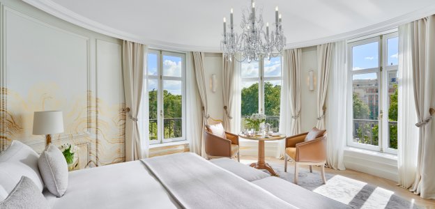 A guest room with a crystal chandelier, a white bed and two leather chairs at a small round table between tall windows overlooking trees.
