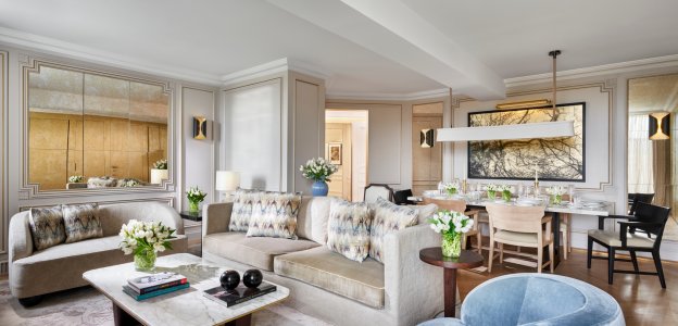 A suite living room with cream sofas around a marble coffee table, a blue velvet chair and a dining table set with flowers beneath a large framed artwork.